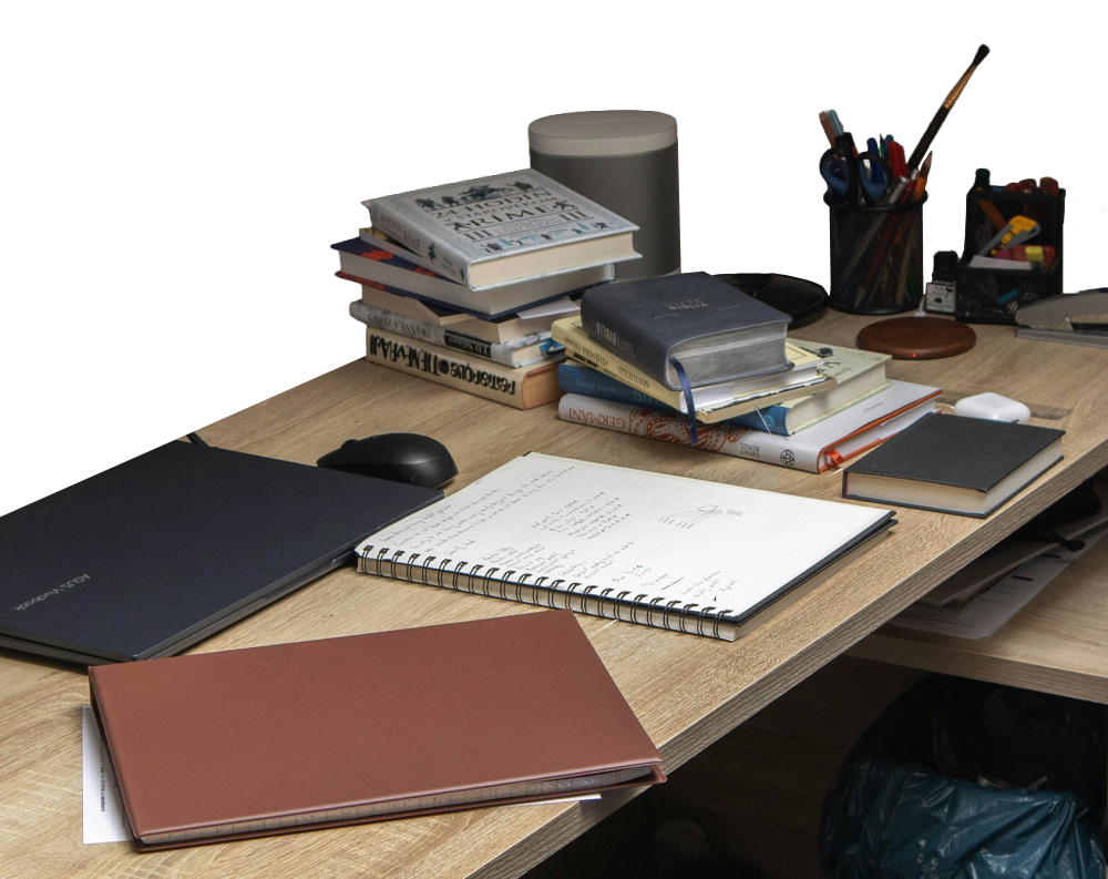 A cluttered wooden desk with stacked books, an open notebook of handwritten notes, and scattered writing tools.