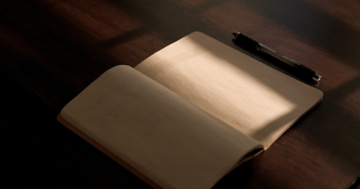 Open notebook on a wooden desk with warm light streaming across blank pages, a pen resting beside it, creating a quiet, reflective mood.